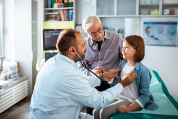 A doctor using a stethoscope to check up on a young patient, who is there with her father.