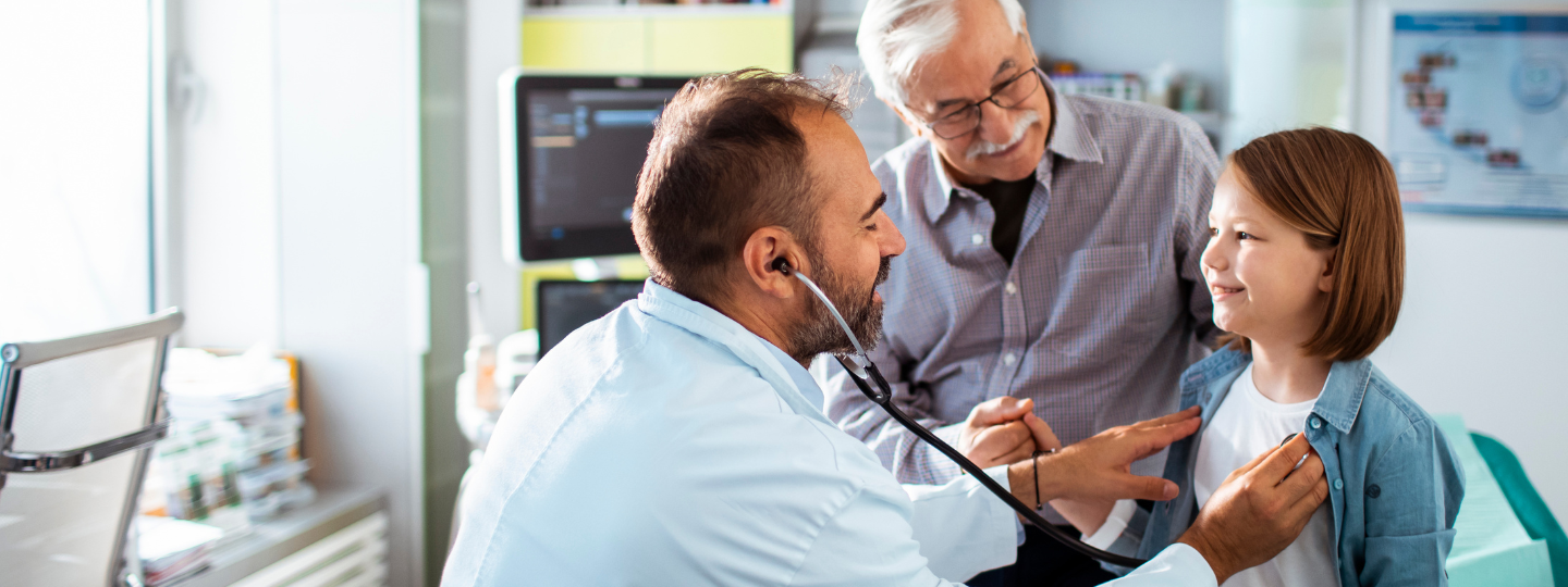 A doctor using a stethoscope to check up on a young patient, who is there with her father.
